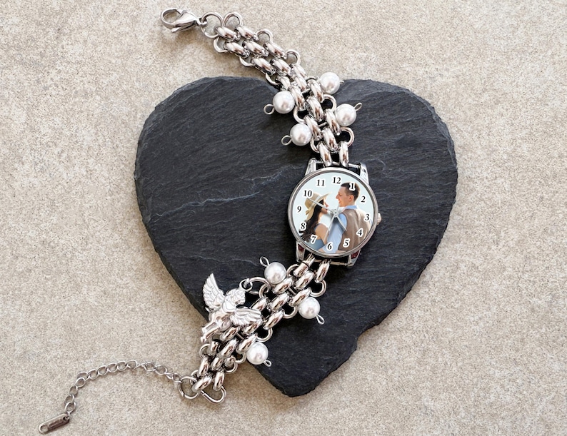 May include: Silver-toned watch bracelet with a chain link design, adorned with faux pearls and a small angel charm. The watch face displays a photo of a couple. The bracelet is resting on a black heart-shaped stone.
