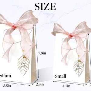 May include: Two white gift bags, one medium and one small, each adorned with a sheer pink ribbon bow. The medium bag is 7.8 inches tall, 5.5 inches wide, and 2.8 inches deep. The small bag is 6.3 inches tall, 4.7 inches wide, and 2.4 inches deep. Each bag has a gold leaf charm.