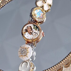 May include: A gold-toned watch with a custom face featuring a person and a dog. The watch is adorned with decorative charms, including a white flower, a starburst, and iridescent accents. The watch is displayed on a reflective surface.