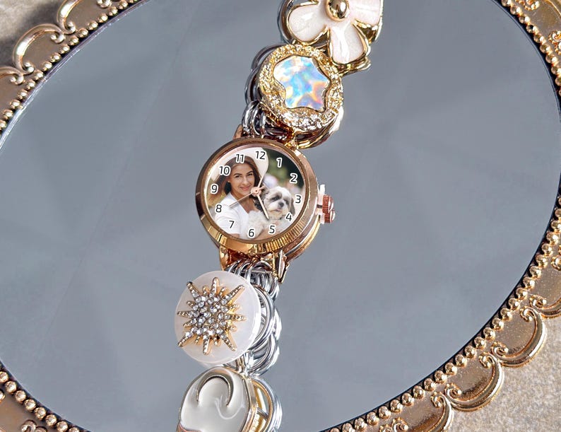 May include: A gold-toned watch with a custom face featuring a person and a dog. The watch is adorned with decorative charms, including a white flower, a starburst, and iridescent accents. The watch is displayed on a reflective surface.