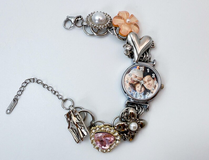 Puede incluir: Pulsera de abalorios plateada con una esfera de reloj que muestra una foto familiar. La pulsera incluye varios abalorios: un coraz&oacute;n, flores, una gema rosa y una perla. La esfera del reloj muestra la hora con n&uacute;meros negros.