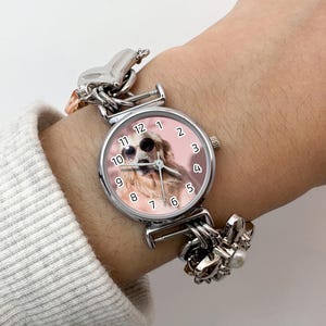 Puede incluir: Un reloj de pulsera plateado con una esfera redonda que muestra un perro con gafas de sol. El reloj tiene un fondo rosa y una correa plateada con abalorios decorativos. El reloj se lleva en una mu&ntilde;eca con una manga de color claro.