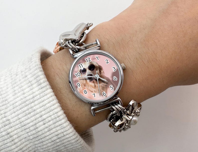 Puede incluir: Un reloj de pulsera plateado con una esfera redonda que muestra un perro con gafas de sol. El reloj tiene un fondo rosa y una correa plateada con abalorios decorativos. El reloj se lleva en una mu&ntilde;eca con una manga de color claro.