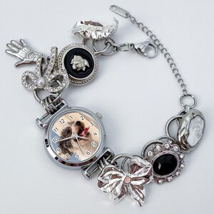 May include: A silver charm bracelet featuring a watch with a dog image, a bow, and various charms. The bracelet includes a hand, a black cameo, and a decorative pendant. The watch face displays the time with silver hands and numbers.
