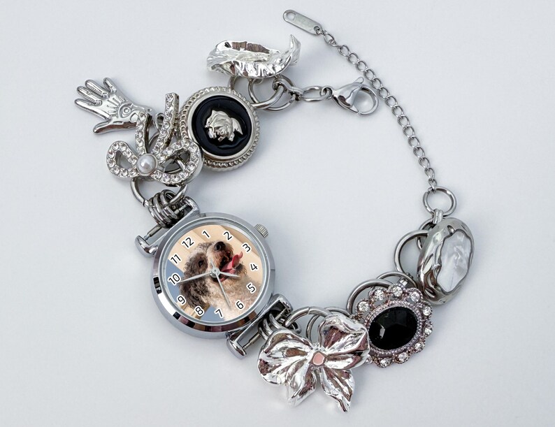 May include: A silver charm bracelet featuring a watch with a dog image, a bow, and various charms. The bracelet includes a hand, a black cameo, and a decorative pendant. The watch face displays the time with silver hands and numbers.