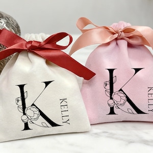 May include: Two small drawstring bags, one off-white and one pink, are tied with satin ribbons. Each bag features a large black letter "K" with floral accents and the name "KELLY" printed vertically.
