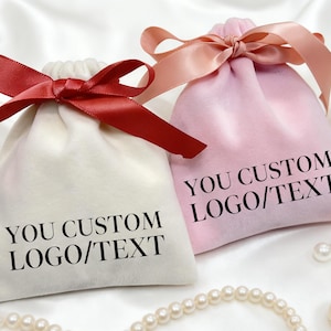 May include: Two velvet drawstring bags, one ivory and one pink, are displayed on a white satin surface. The ivory bag has a red satin ribbon, and the pink bag has a peach-coloured ribbon. Both bags have the text "YOU CUSTOM LOGO/TEXT" printed on them.