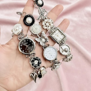 May include: Three silver-toned watches with decorative embellishments. One watch has a black face, another a white face, and the third a rectangular face. Embellishments include pearls, flowers, and a butterfly. The watches are displayed on a pink background.