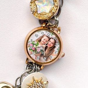 May include: A rose gold watch with a photo of two people on the face. The watch is part of a charm bracelet with gold, silver, and white decorative elements. The watch face has black numbers and hands.