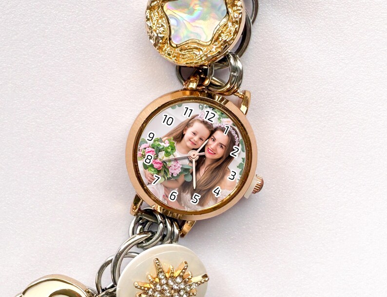 May include: A rose gold watch with a photo of two people on the face. The watch is part of a charm bracelet with gold, silver, and white decorative elements. The watch face has black numbers and hands.