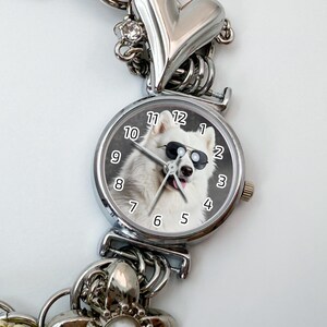 Puede incluir: Reloj plateado con un perro blanco con gafas de sol en la esfera. El reloj tiene una correa plateada con detalles de coraz&oacute;n y abalorios. La esfera muestra los n&uacute;meros del 1 al 12.