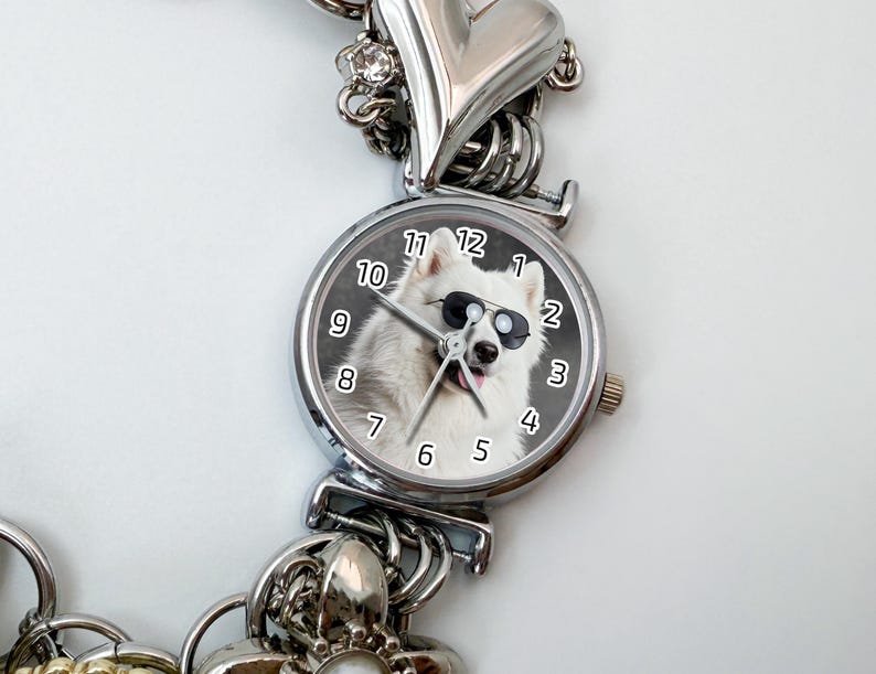 Puede incluir: Reloj plateado con un perro blanco con gafas de sol en la esfera. El reloj tiene una correa plateada con detalles de coraz&oacute;n y abalorios. La esfera muestra los n&uacute;meros del 1 al 12.