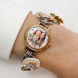 May include: A gold-toned watch with a round face featuring a photo of a woman and a child. The watch has a gold bezel, a white dial with black numerals, and a linked bracelet with decorative charms. The watch is worn on a wrist.