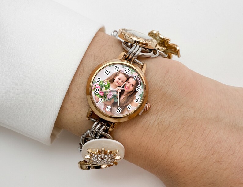 May include: A gold-toned watch with a round face featuring a photo of a woman and a child. The watch has a gold bezel, a white dial with black numerals, and a linked bracelet with decorative charms. The watch is worn on a wrist.