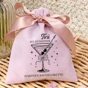Puede incluir: Una bolsa de terciopelo rosa claro con cierre de cordón y lazo de satén. La bolsa presenta un gráfico negro de una copa de martini con aceitunas y el texto "Tini Bit Hungover" y "Harper's Bachelorette".