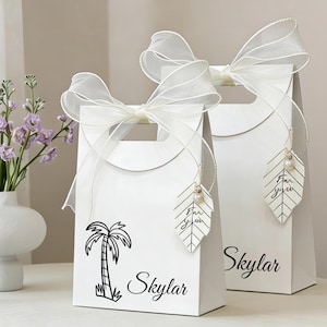 May include: Two white gift bags with a palm tree design and the name "Skylar" printed in black. Each bag has a white ribbon bow and a leaf-shaped tag that says "For You".