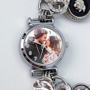 May include: Silver-toned watch with a round face featuring a couple kissing. The watch face has black numerals and hands. The watch is part of a bracelet with decorative charms, including a heart and a circular charm with rhinestones.