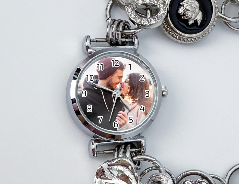 May include: Silver-toned watch with a round face featuring a couple kissing. The watch face has black numerals and hands. The watch is part of a bracelet with decorative charms, including a heart and a circular charm with rhinestones.