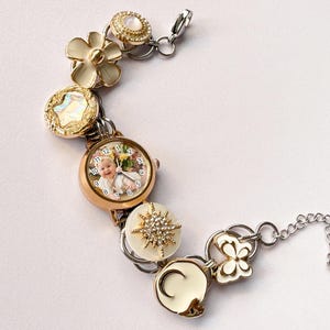 May include: A gold-toned bracelet featuring a watch face with a baby's photo, surrounded by decorative elements. The bracelet includes flower, starburst, and butterfly charms, with a silver chain and clasp. The bracelet is approximately 7 inches long.
