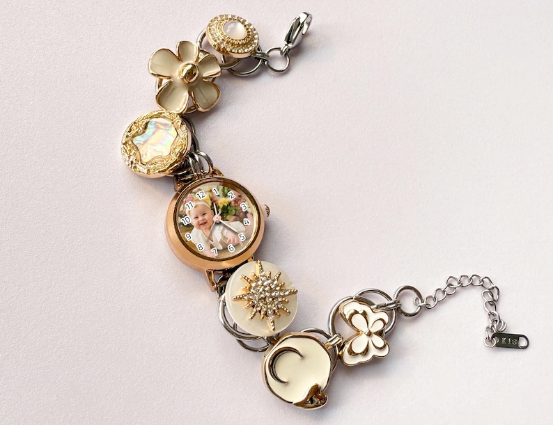 May include: A gold-toned bracelet featuring a watch face with a baby's photo, surrounded by decorative elements. The bracelet includes flower, starburst, and butterfly charms, with a silver chain and clasp. The bracelet is approximately 7 inches long.