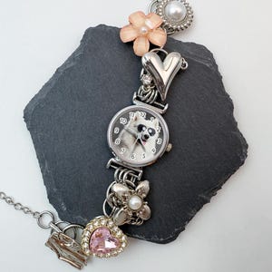Puede incluir: Reloj plateado con esfera redonda que muestra un perro con gafas de sol. La correa del reloj est&aacute; adornada con abalorios, incluyendo una flor rosa, un coraz&oacute;n y una joya en forma de coraz&oacute;n rosa. Una cadena plateada se extiende desde el reloj.