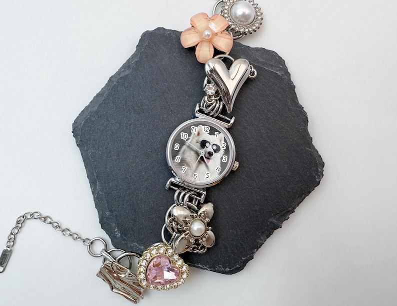 Puede incluir: Reloj plateado con esfera redonda que muestra un perro con gafas de sol. La correa del reloj est&aacute; adornada con abalorios, incluyendo una flor rosa, un coraz&oacute;n y una joya en forma de coraz&oacute;n rosa. Una cadena plateada se extiende desde el reloj.