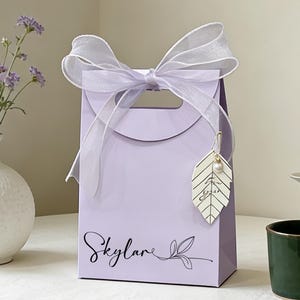 May include: A lavender gift bag with a white ribbon bow and a leaf-shaped tag. The bag has the name "Skylare" in script. A white vase with purple flowers and a green mug are also in the image.