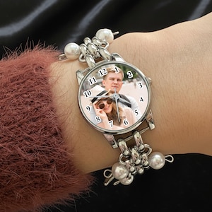 May include: Silver wristwatch with a round face featuring a photo of a couple. The watch has a silver band with pearl accents. The watch is being worn on a wrist, with a maroon sweater sleeve visible.