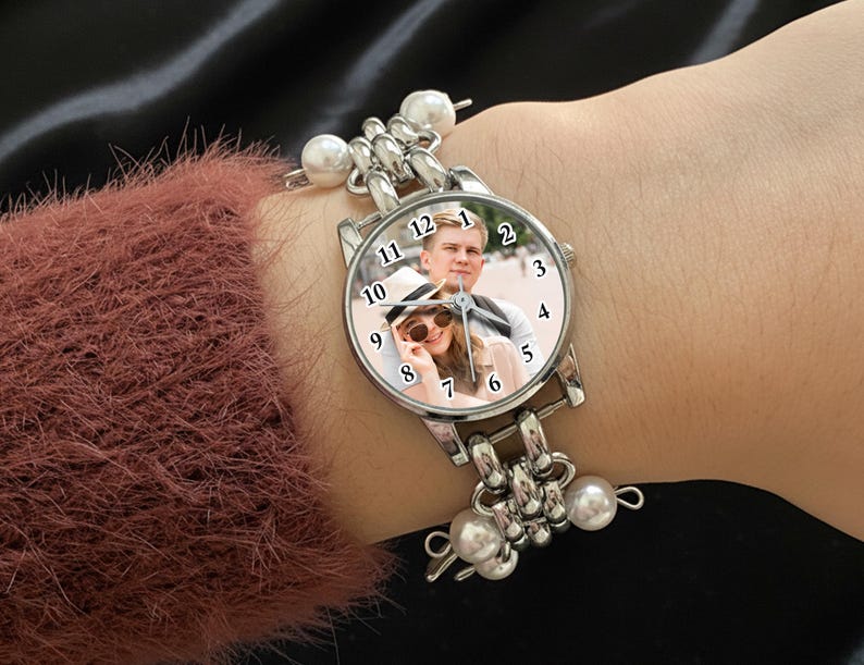 May include: Silver wristwatch with a round face featuring a photo of a couple. The watch has a silver band with pearl accents. The watch is being worn on a wrist, with a maroon sweater sleeve visible.