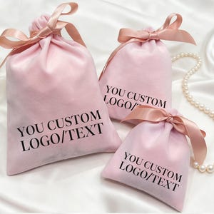 Könnte beinhalten: Drei rosa Samt-Kordelzugbeutel mit Satinbändern. Die Beutel tragen den Text "YOU CUSTOM LOGO/TEXT" in Schwarz. Eine Perlenkette und weiße Blumen sind im Hintergrund.