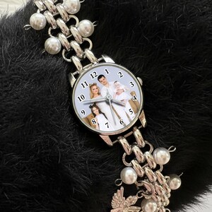 May include: Silver-toned wristwatch with a family photo on the watch face. The watch has a chain-link band with pearl accents. The watch is displayed on a black, textured surface.