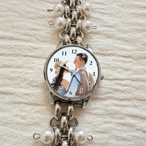 May include: A silver-toned wristwatch with a round face featuring a couple's photo. The watch has a linked band with faux pearl accents. The watch face displays the numbers 1 through 12. The watch is on a white textured surface.