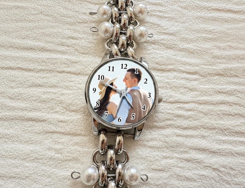 May include: A silver-toned wristwatch with a round face featuring a couple's photo. The watch has a linked band with faux pearl accents. The watch face displays the numbers 1 through 12. The watch is on a white textured surface.