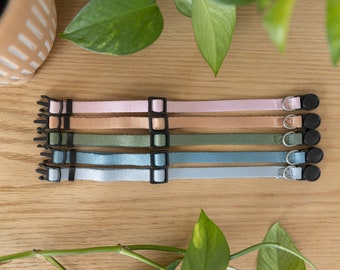 May include: A collection of six pastel-colored cat collars with black plastic buckles and metal D-rings. The collars are arranged on a light wood surface, with green plants in the background. Colors include pink, peach, green, and blue.
