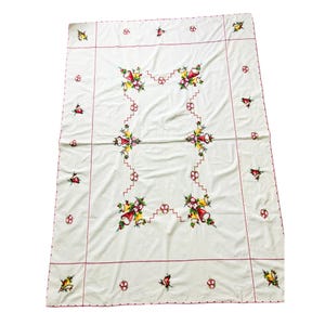 May include: A white rectangular tablecloth featuring embroidered red and yellow bell motifs with green holly leaves. The design includes a central zigzag border and a thin red stitched frame around the perimeter with scalloped edges.