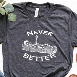 May include: A dark gray t-shirt with a white graphic of a skeleton lying in a coffin with the text "Never Better".