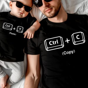 May include: Black t-shirts with white graphic keyboard symbols. One shirt has "Ctrl + C (Copy)" and the other has "Ctrl + V (Paste)". The shirts are worn by a parent and child.
