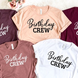 May include: A collection of five t-shirts in various colors: burgundy, peach, mauve, white, and maroon. Each shirt features the words "Birthday Crew" in a black, cursive font. The shirts are neatly arranged on a white surface.