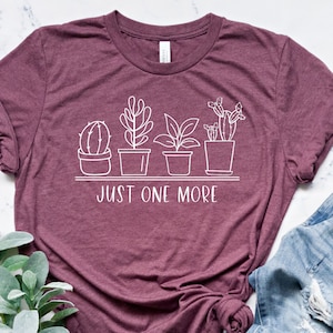 May include: Burgundy t-shirt with a white line drawing of potted plants and the text "JUST ONE MORE". The shirt is paired with blue jeans and a sprig of greenery.