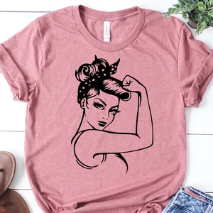 Może przedstawiać: R&oacute;żowy T-shirt z czarną sylwetką kobiety z podniesioną pięścią. Kobieta ma w włosach bandanę w kropki.