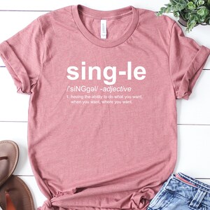 May include: A dusty rose t-shirt with the word "single" in large white letters. Below the word is a definition, including the pronunciation and the phrase "having the ability to do what you want, when you want, where you want."