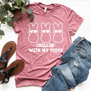 Peut inclure: Un t-shirt rose avec un motif blanc de trois Peeps de P&acirc;ques portant des lunettes de soleil. Le texte "Chillin' with my peeps" est imprim&eacute; sous les Peeps.