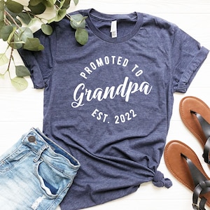 May include: A navy blue t-shirt with white text that reads "Promoted to Grandpa Est. 2022".