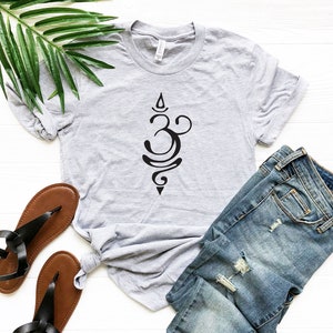 May include: A gray t-shirt with a black Om symbol design. The shirt is laid out on a white surface with a pair of brown sandals and blue jeans.