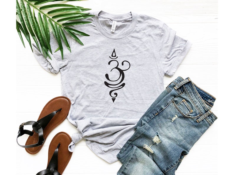 May include: A gray t-shirt with a black Om symbol design. The shirt is laid out on a white surface with a pair of brown sandals and blue jeans.
