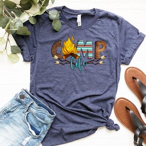 May include: A navy blue t-shirt featuring a graphic design of the word "camp" with a campfire and marshmallows. The word "life" is written in teal below the word "camp".
