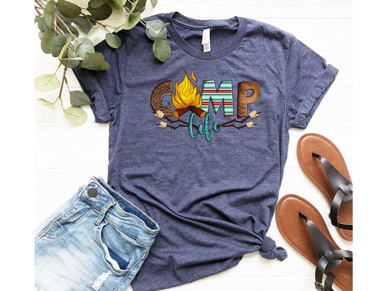 May include: A navy blue t-shirt featuring a graphic design of the word "camp" with a campfire and marshmallows. The word "life" is written in teal below the word "camp".