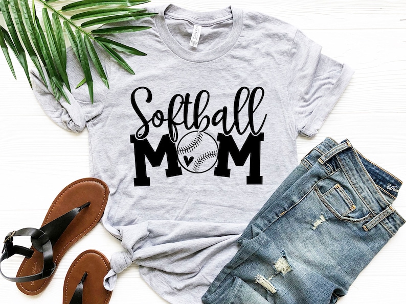 Puede incluir: Una camiseta gris con el texto "Softball Mom" en letras negras y un gr&aacute;fico de softball negro con un peque&ntilde;o coraz&oacute;n.