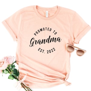 May include: A peach-coloured t-shirt with the text "Promoted to Grandma Est. 2022" in black lettering.