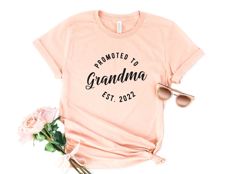 May include: A peach-coloured t-shirt with the text "Promoted to Grandma Est. 2022" in black lettering.